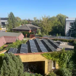 Panele fotowoltaiczne na dachu garażu, widok z góry, otoczone zielenią. Słoneczny dzień, w tle domy i drzewa. Nietypowy montaż paneli.