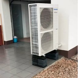 Atom Sp. z o.o. - Biała, dwuczęściowa klimatyzacja Panasonic Inverter, zamontowana na czarnych cegłach przy ścianie budynku z szarym chodnikiem i kamienistym podłożem.