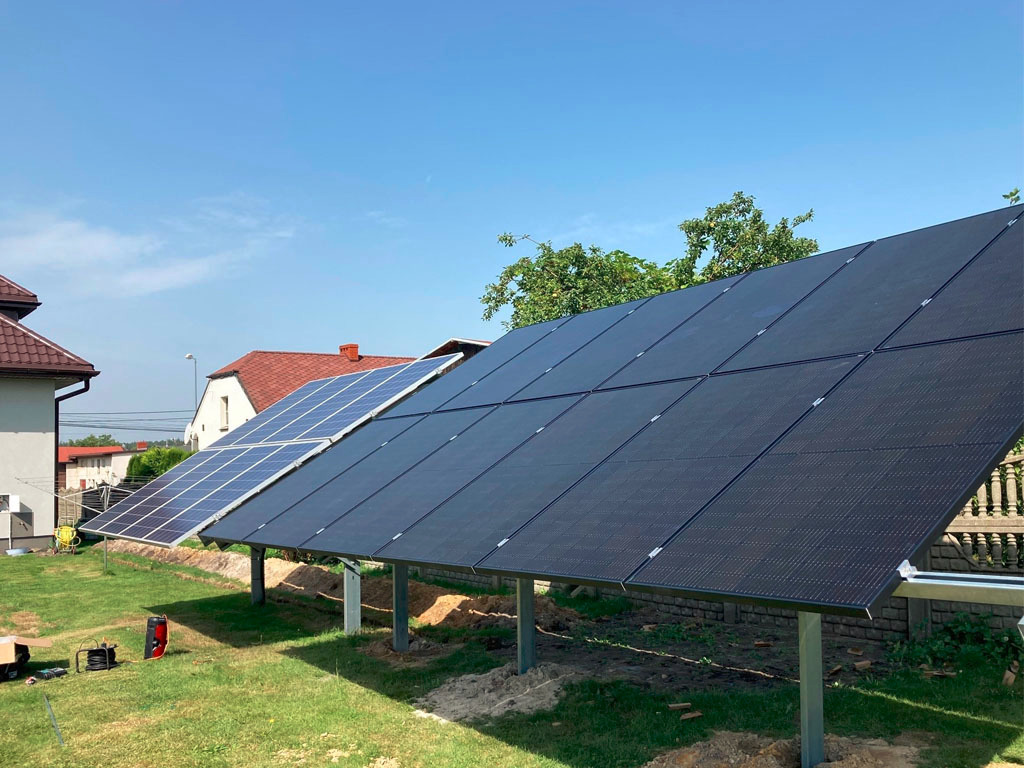 Instalacja paneli fotowoltaicznych na trawniku przed domem. Widoczne dwa rzędy paneli: jeden z czarnymi panelami, drugi z niebieskimi. Błękitne niebo w tle.