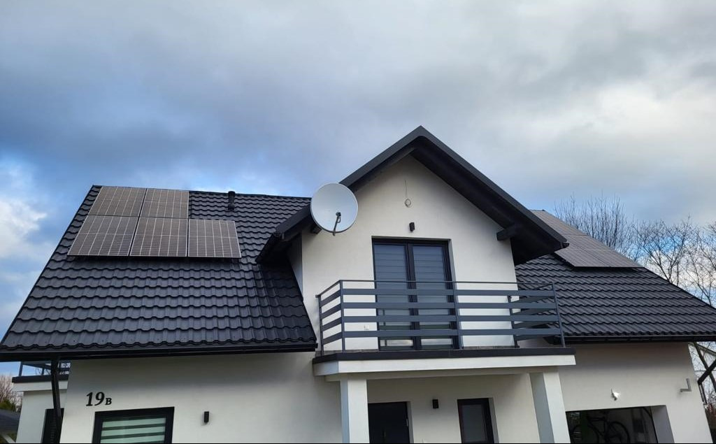 Biały dom z ciemnym dachem, na którym zamontowano panele fotowoltaiczne. Widoczna antena satelitarna i numer domu 19b. Pochmurne niebo w tle.
