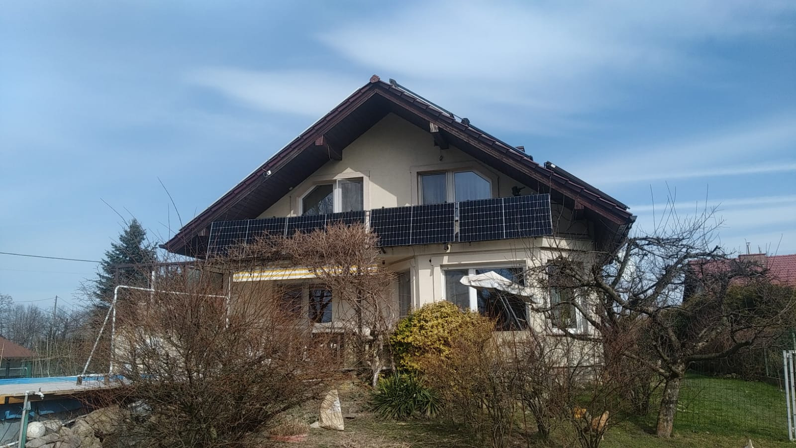 Dwupiętrowy dom z panelami fotowoltaicznymi zamontowanymi na balkonie. Widoczne drzewa bez liści i błękitne niebo.
