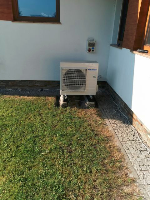 Zewnętrzna jednostka klimatyzacji Panasonic na trawniku przy ścianie domu z oknami. Widoczna skrzynka elektryczna na ścianie. Instalacja klimatyzacji.