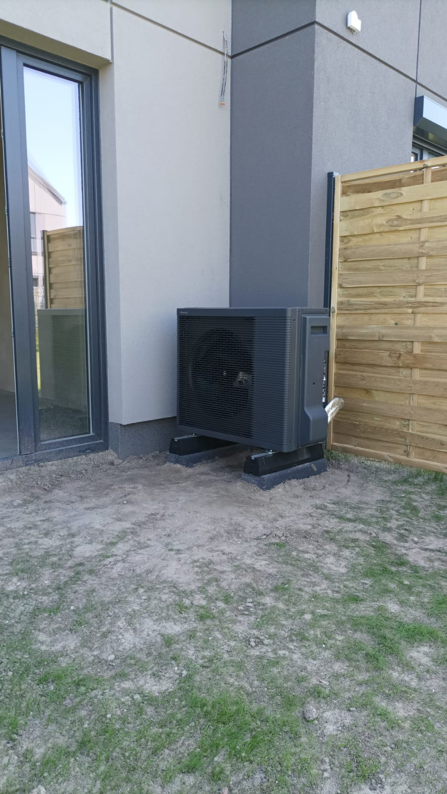 Czarna pompa ciepła Panasonic Aquarea zamontowana na betonowych podstawach przy nowoczesnym domu z szarą stolarką okienną i drewnianym płotem.