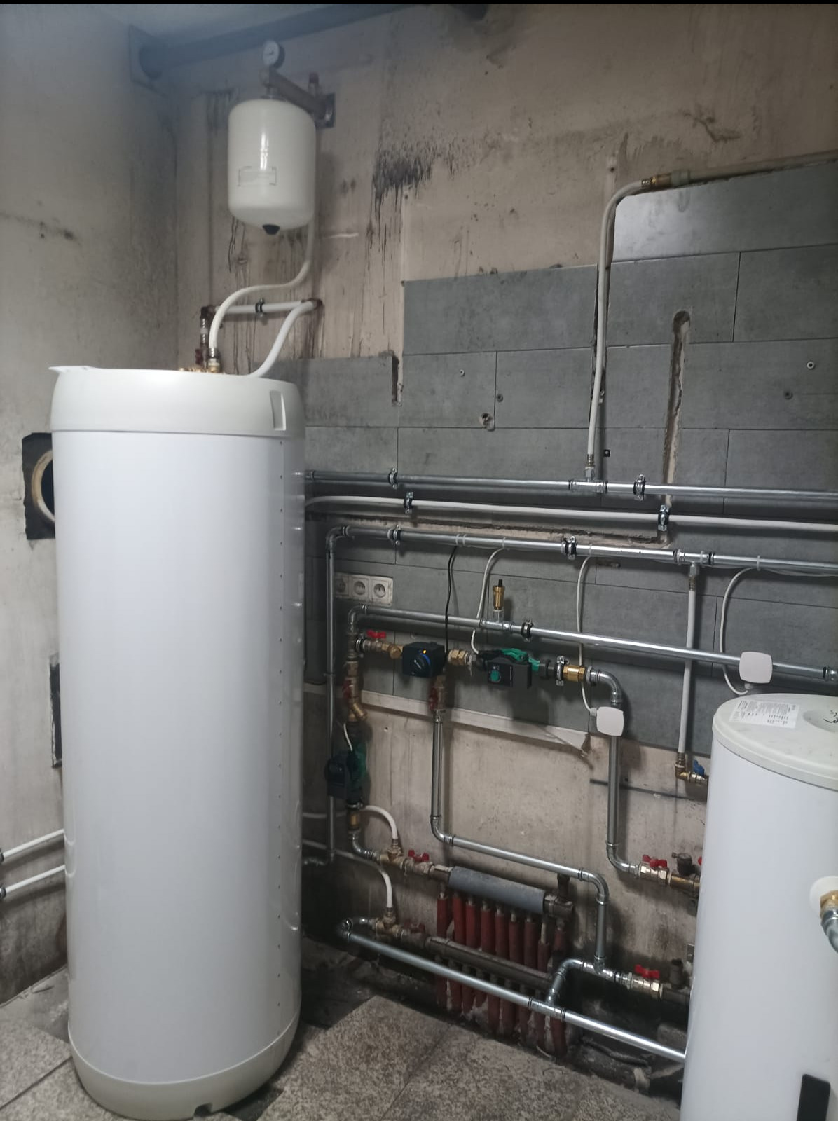 Biały zbiornik akumulacyjny ciepła w piwnicy, z widoczną instalacją rurową i grzejnikiem. Szare płytki częściowo zakrywają ścianę.