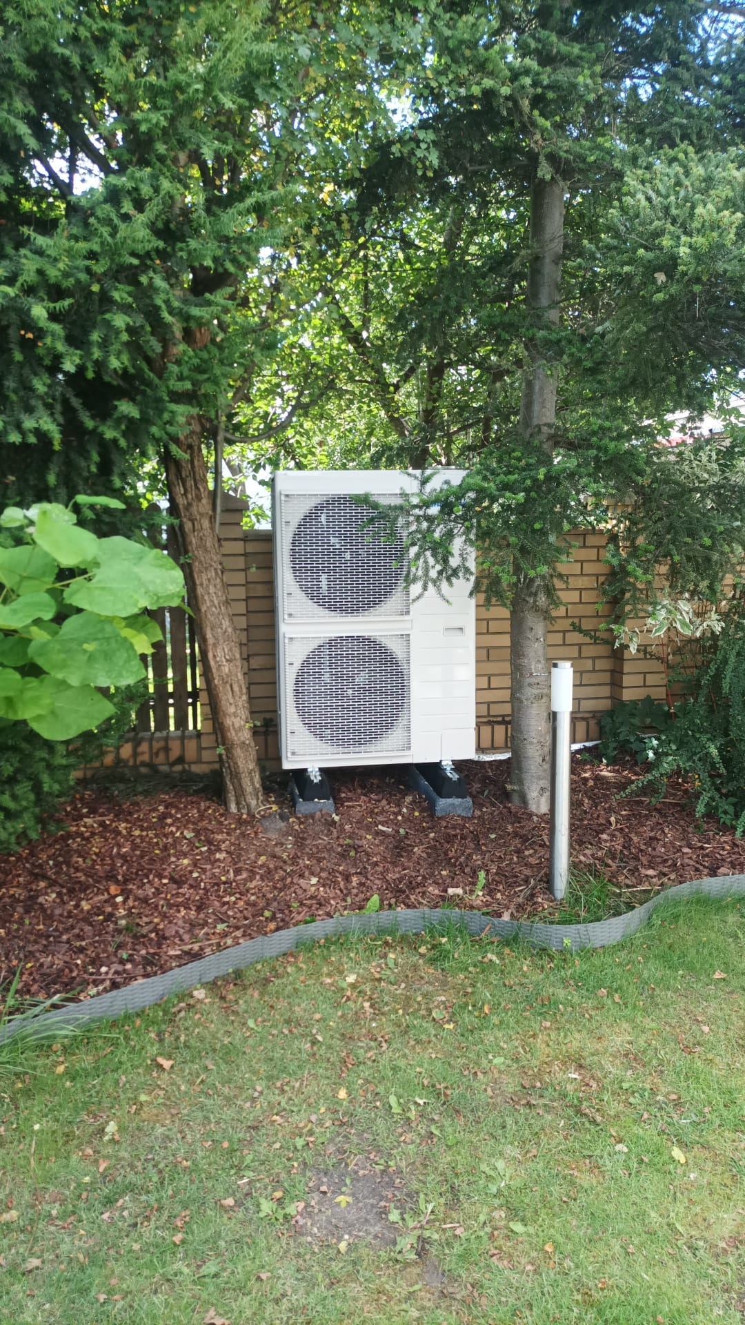 Biała, dwuczęściowa pompa ciepła na betonowych podstawach, stoi na korze drzewnej, otoczona zielenią i ceglanym murem. Widok z poziomu trawnika.