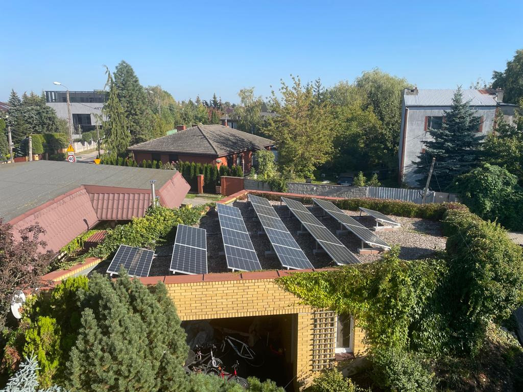 Panele fotowoltaiczne na dachu garażu, widok z góry, otoczone zielenią. Słoneczny dzień, w tle domy i drzewa. Nietypowy montaż paneli.