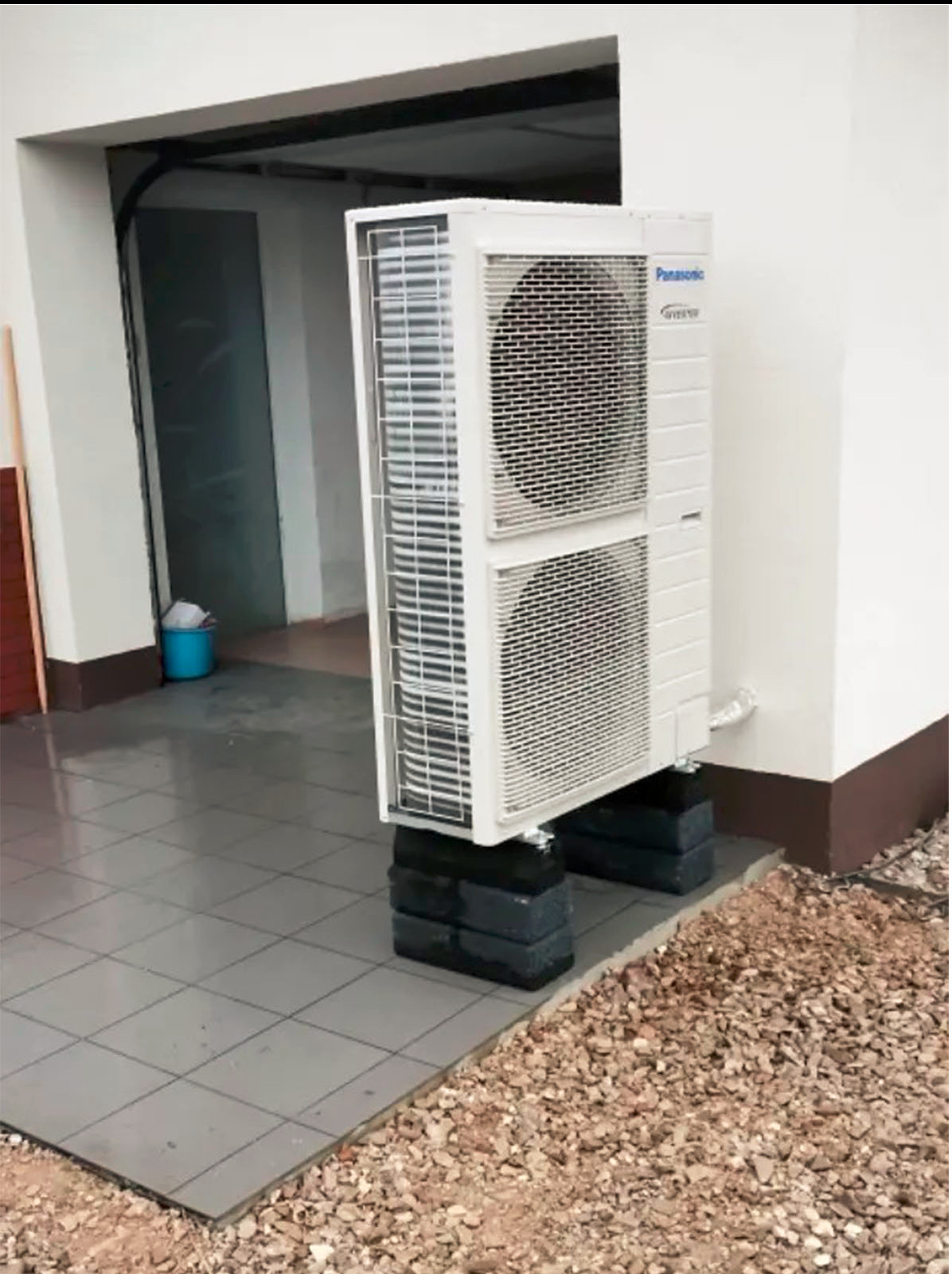 Biała, dwuczęściowa klimatyzacja Panasonic Inverter, zamontowana na czarnych cegłach przy ścianie budynku z szarym chodnikiem i kamienistym podłożem.