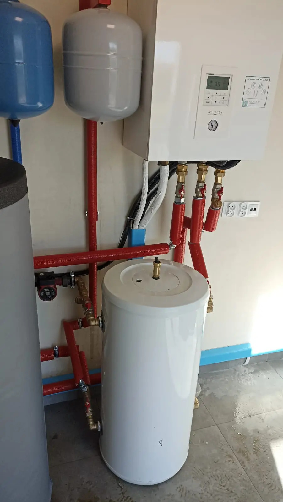 Zestaw urządzeń, w tym biały zbiornik i panel sterowania Aquarea Smart Cloud, z czerwonymi rurami i niebieskimi zbiornikami ciśnieniowymi na kremowej ścianie.