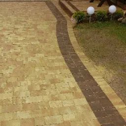 Ułożona nawierzchnia z kostki brukowej w odcieniach beżu i brązu, z dekoracyjnym pasem ciemniejszej kostki biegnącym po łuku, prowadzącym do schodów i ogrodu z elementami oświetlenia.