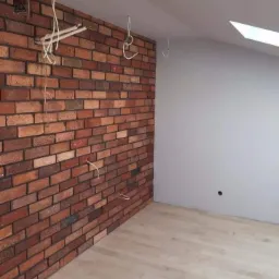 Wnętrze w Kartuzach w trakcie prac wykończeniowych: ściana z cegły klinkierowej, fragment ściany pomalowany na szaro, jasna podłoga z paneli, widoczne kable i otwory instalacyjne.