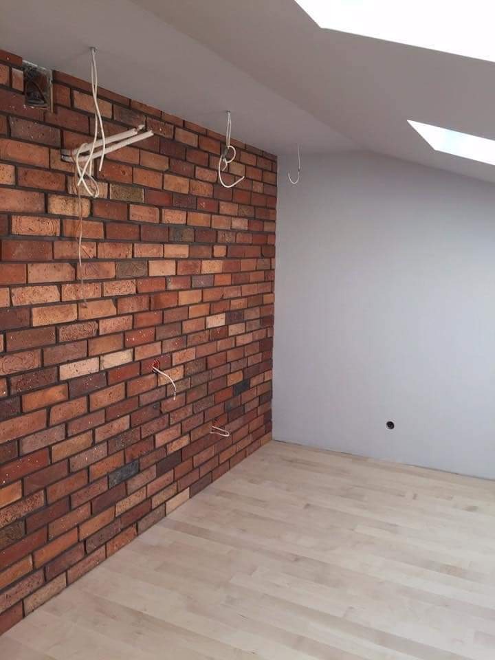 Wnętrze w Kartuzach w trakcie prac wykończeniowych: ściana z cegły klinkierowej, fragment ściany pomalowany na szaro, jasna podłoga z paneli, widoczne kable i otwory instalacyjne.