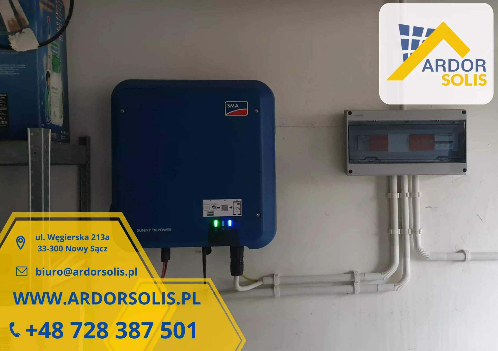 Niebieski falownik SMA Sunny Tripower zamontowany na białej ścianie obok skrzynki elektrycznej z białymi rurami kablowymi.