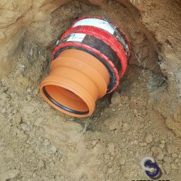 Instal - Gaz Milena Szurlej - Pomarańczowa rura gazowa w wykopie, z czarną uszczelką i czerwoną izolacją termiczną, zabezpieczona metalowymi obejmami, widoczna ziemia i logo firmy instalacyjnej.