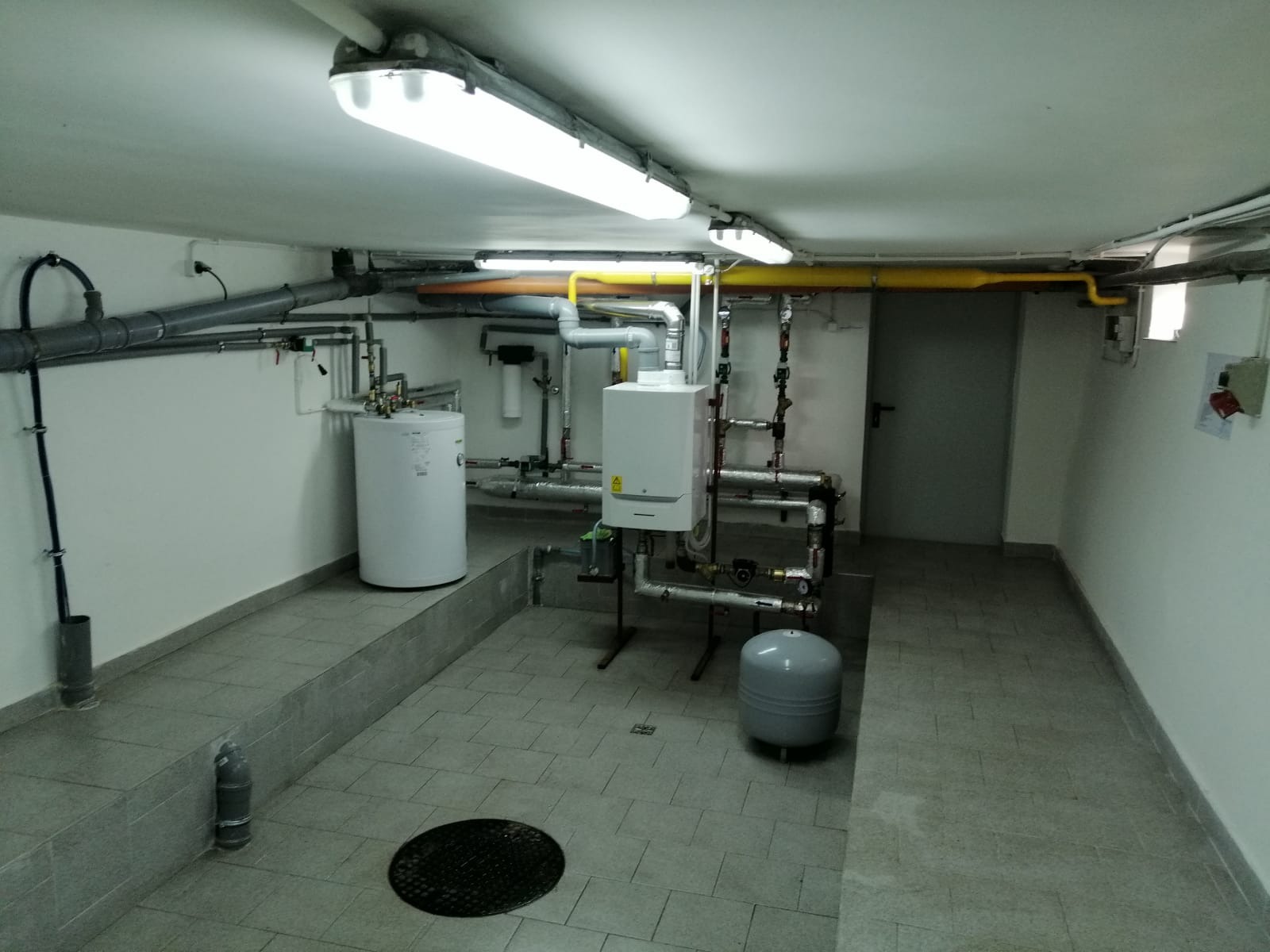 Kotłownia z białym kotłem gazowym, zbiornikiem na wodę i szarym zbiornikiem wyrównawczym, widoczne rury instalacji grzewczej i kanalizacyjnej pod sufitem, szara podłoga z kratką ściekową.