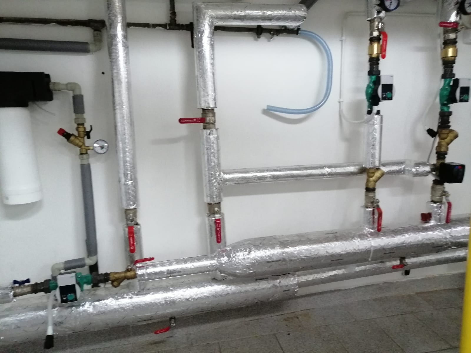 Izolowane rury instalacji grzewczej z zaworami i manometrami na tle białej ściany, widoczne także elementy filtra wody.