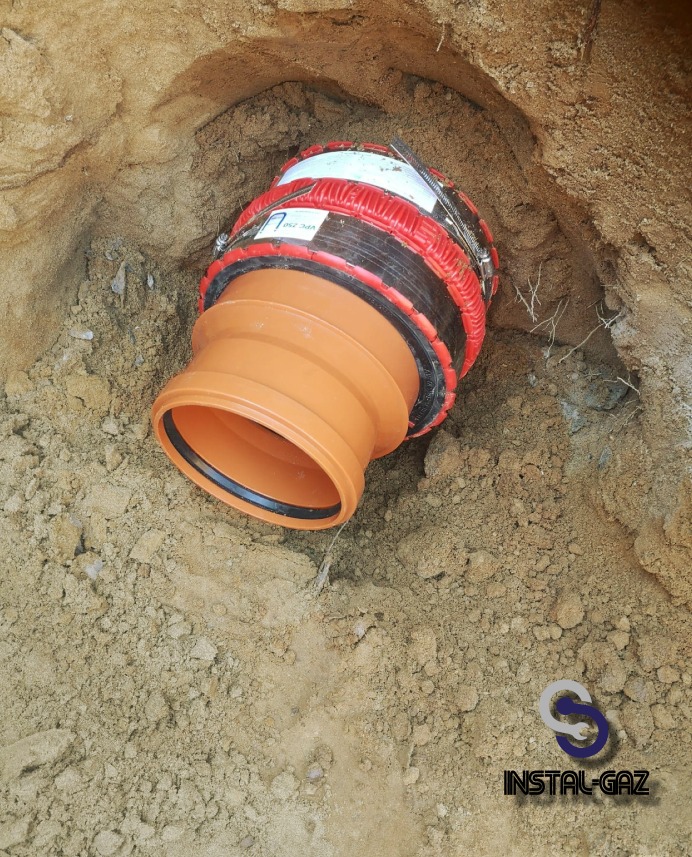 Pomarańczowa rura gazowa w wykopie, z czarną uszczelką i czerwoną izolacją termiczną, zabezpieczona metalowymi obejmami, widoczna ziemia i logo firmy instalacyjnej.