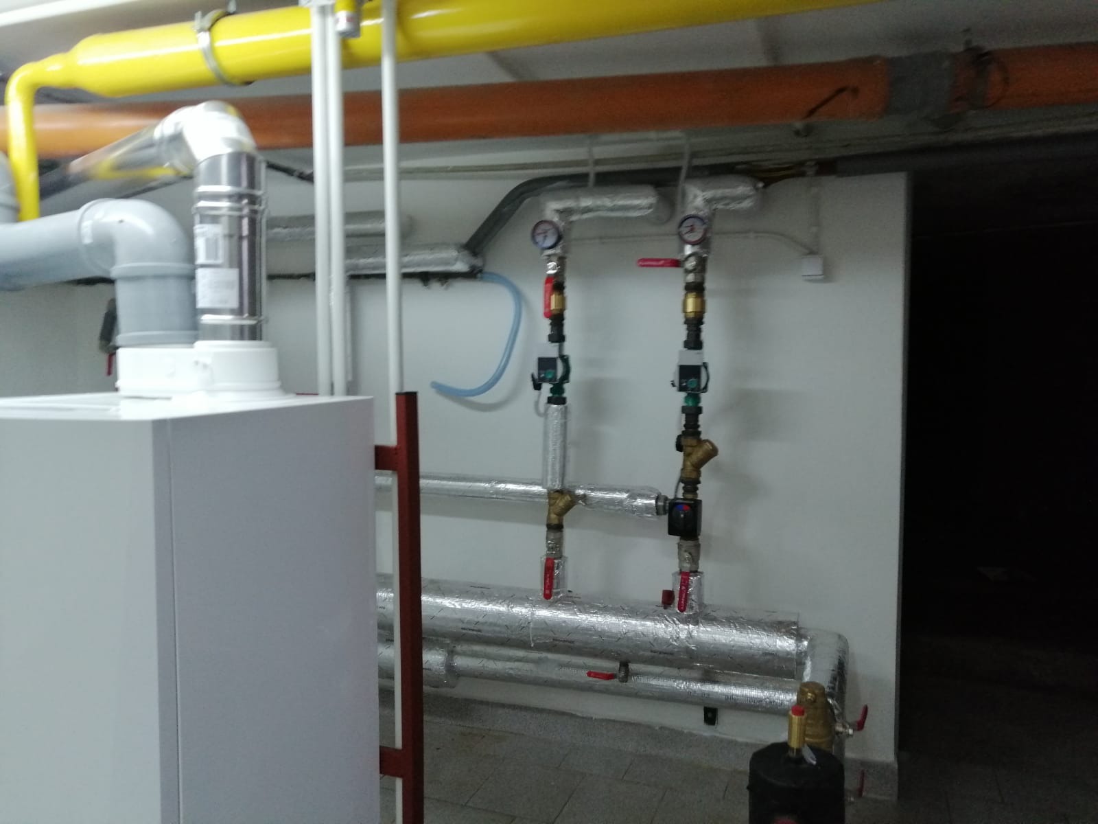 Biały piec grzewczy z kominem, rury w izolacji aluminiowej z zaworami i manometrami na ścianie, żółta rura gazowa pod sufitem.