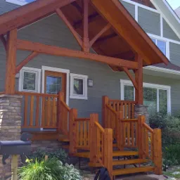 Zadaszone wejście do domu z drewnianą konstrukcją, kamiennymi filarami i drewnianymi schodami z balustradą, świeżo po impregnacji i zabezpieczeniu drewna.