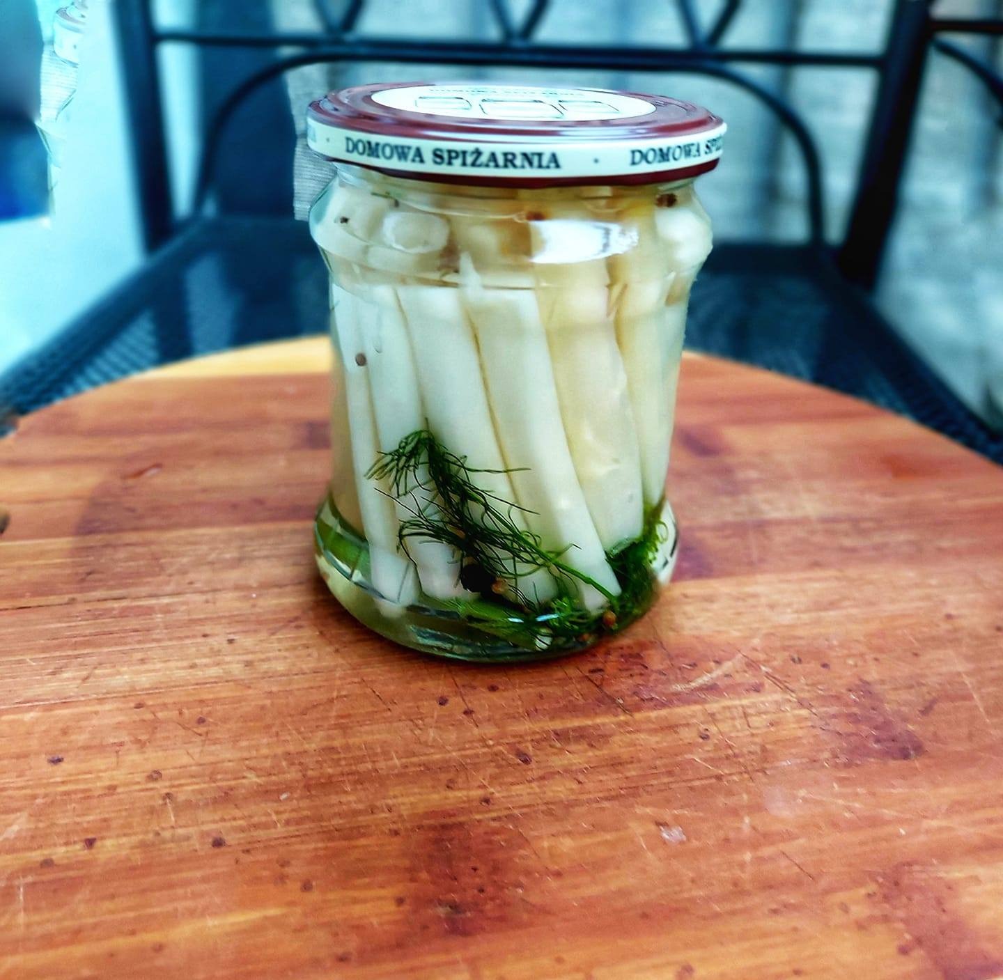 Słoik kiszonych, białych szparagów z koperkiem, zamknięty brązową nakrętką z napisem 'Domowa Spiżarnia', ustawiony na drewnianej desce, z fragmentem krzesła w tle.
