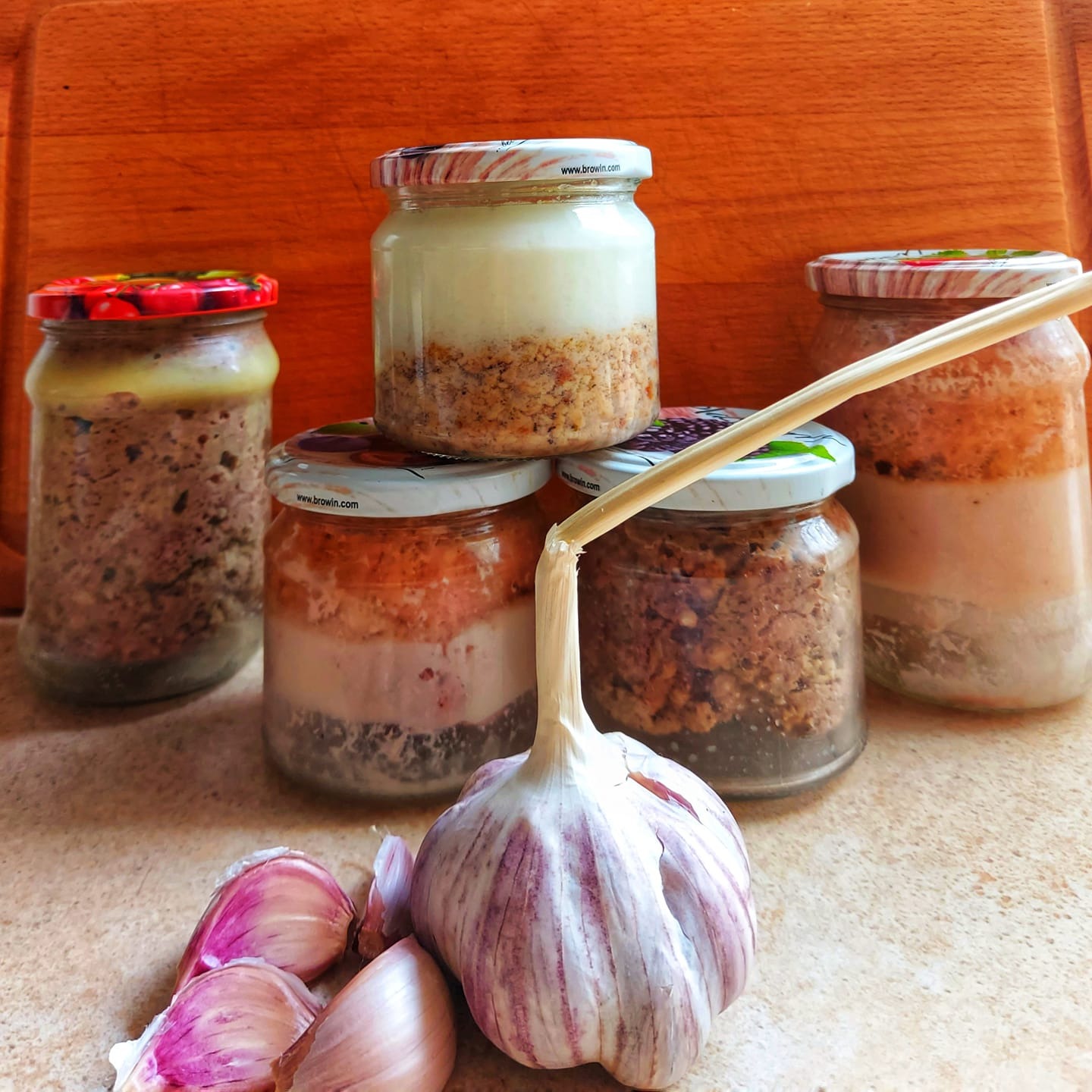 Słoiki z domowymi przetworami mięsnymi i smalcem, ustawione na blacie, z główką czosnku i kilkoma ząbkami na pierwszym planie, na tle drewnianej deski do krojenia.