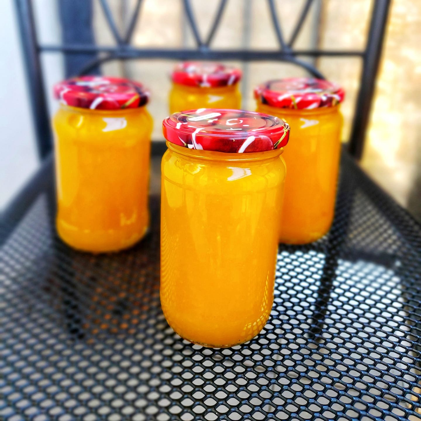 Cztery słoiki marmolady dyniowo-pomarańczowej z czerwonymi, wzorzystymi wieczkami, ustawione na metalowej, ażurowej powierzchni krzesła.