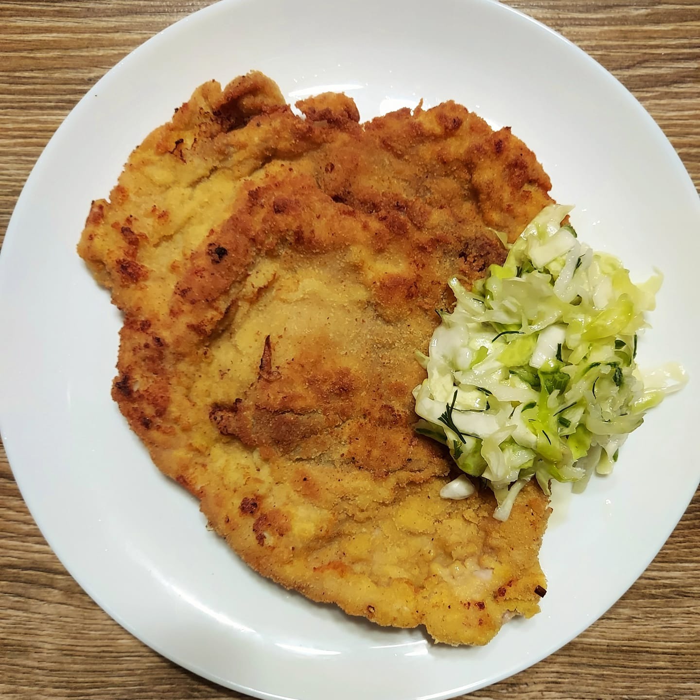 Kawałek panierowanej łopatki wieprzowej na białym talerzu, podany z surówką z kapusty, ogórka i kalarepy, na drewnianym stole.