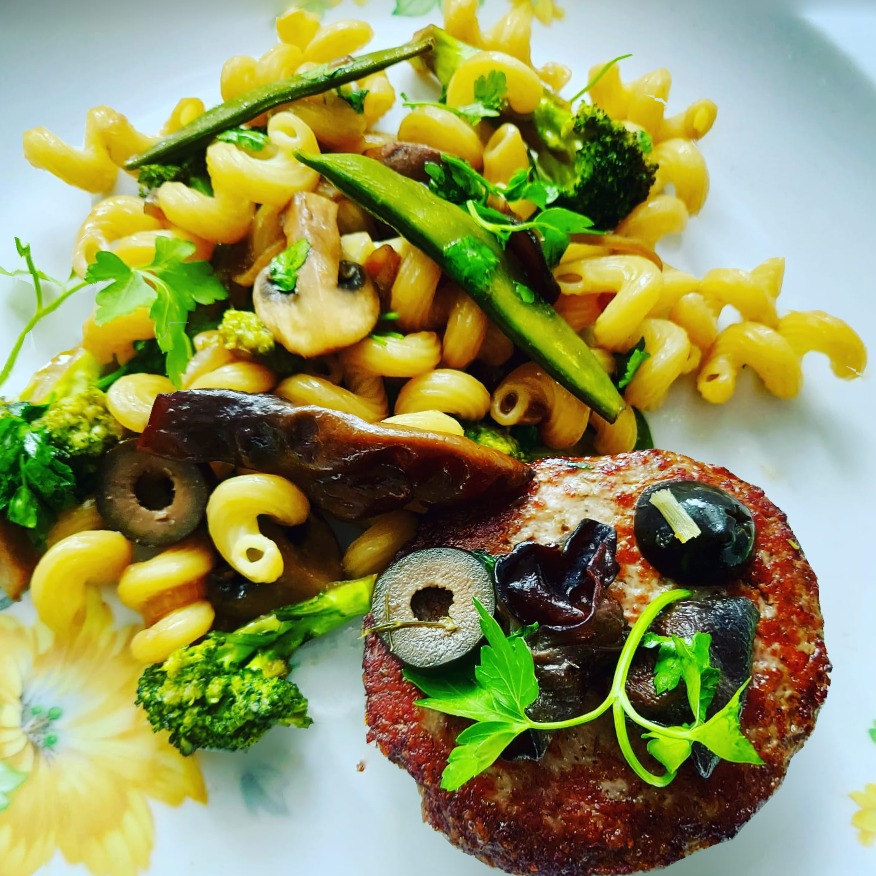 Burger cielęcy podany z makaronem cavatappi, brokułami, groszkiem cukrowym, czarnymi pieczarkami, grzybami mun, oliwkami i pietruszką na talerzu z kwiatowym wzorem.