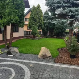 Starannie zaprojektowany ogród przydomowy z równo przystrzyżonym trawnikiem, ozdobnymi kamieniami i wysypaną korą, widoczne drzewa iglaste i kostka brukowa w geometryczny wzór.