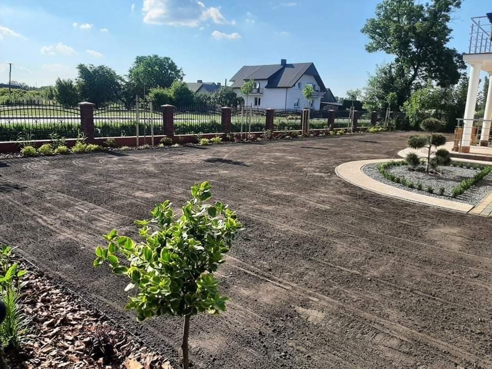 Przygotowany teren pod przyszły ogród z widocznymi śladami po maszynach, świeżo posadzone krzewy i drzewka, w tle dom z ciemnym dachem i ogrodzenie z ceglanymi słupkami.