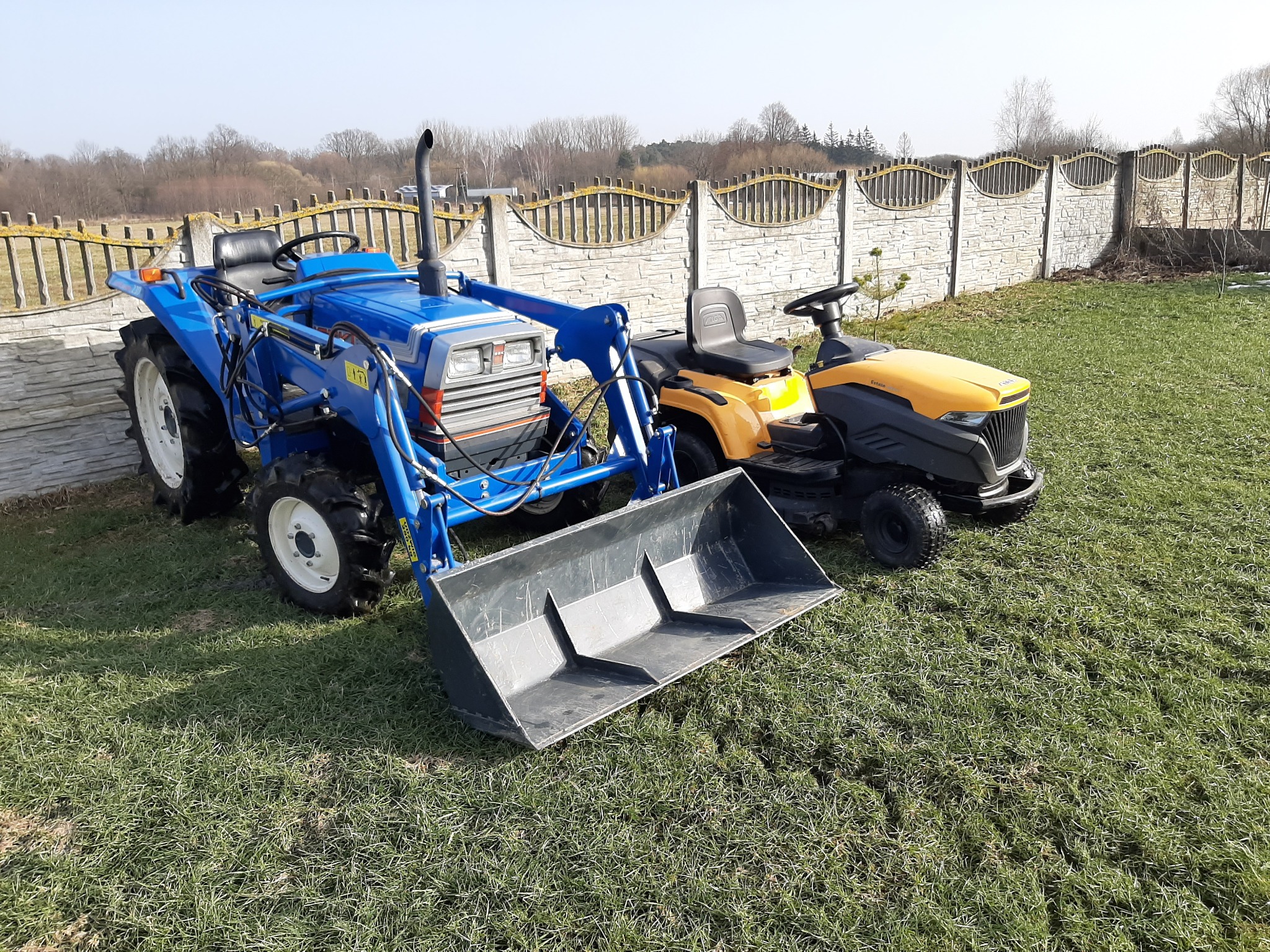 Niebieski mini-traktor z ładowarką czołową i żółta kosiarka samojezdna stoją na trawniku przed betonowym ogrodzeniem z ozdobnymi przęsłami.