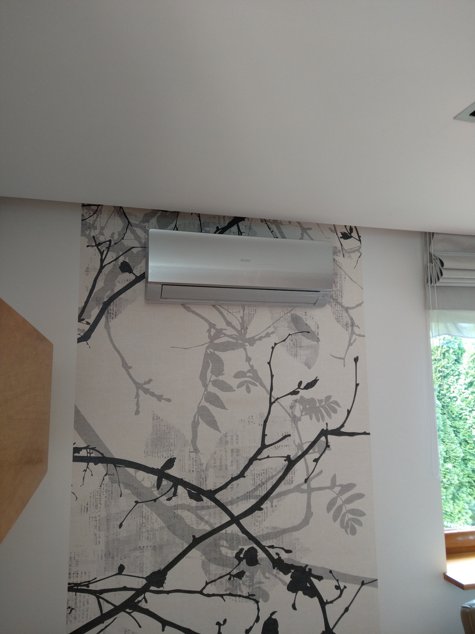 Biała jednostka klimatyzacji marki Haier zamontowana na ścianie z tapetą w motywie gałęzi drzew, widok z dołu.