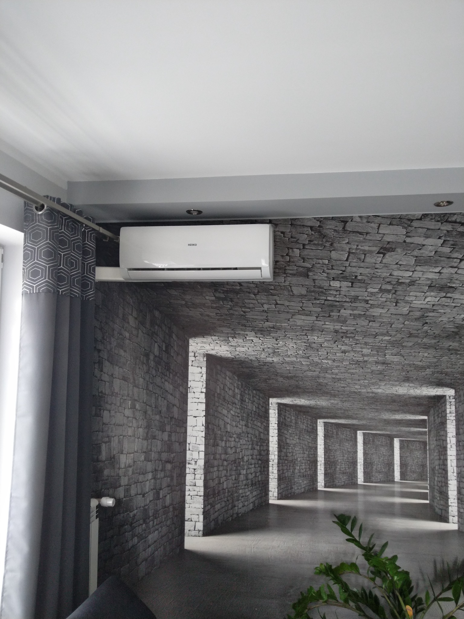 Klimatyzator marki Heiko zamontowany w pomieszczeniu we Wrzeczku z fototapetą imitującą kamienny tunel, widok z dołu, zasłony w geometryczne wzory.
