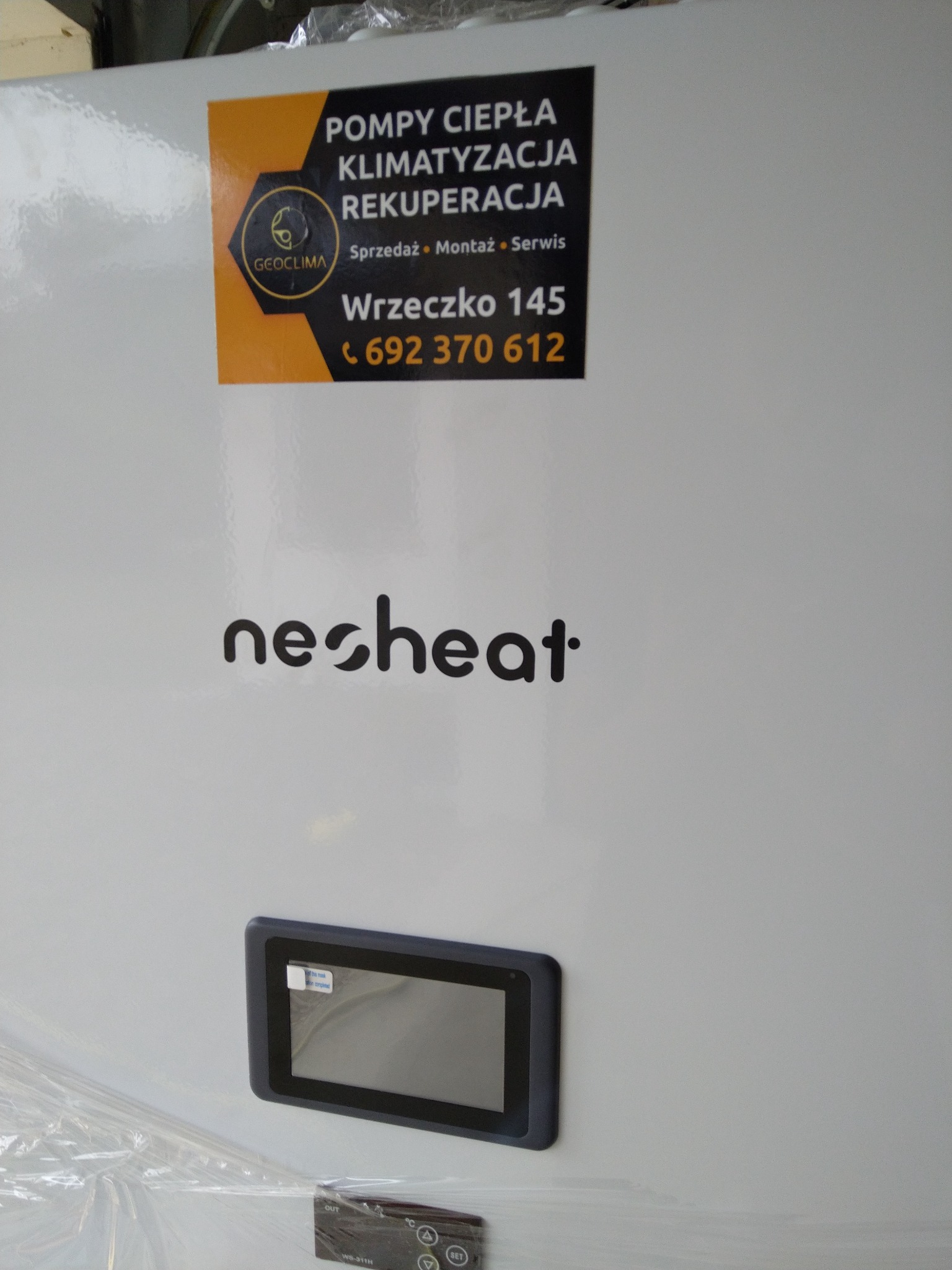 Biała pompa ciepła marki nesheat z panelem sterowania i naklejką informacyjną firmy GEOCLIMA oferującej sprzedaż, montaż i serwis pomp ciepła, klimatyzacji i rekuperacji, z widocznym adresem...