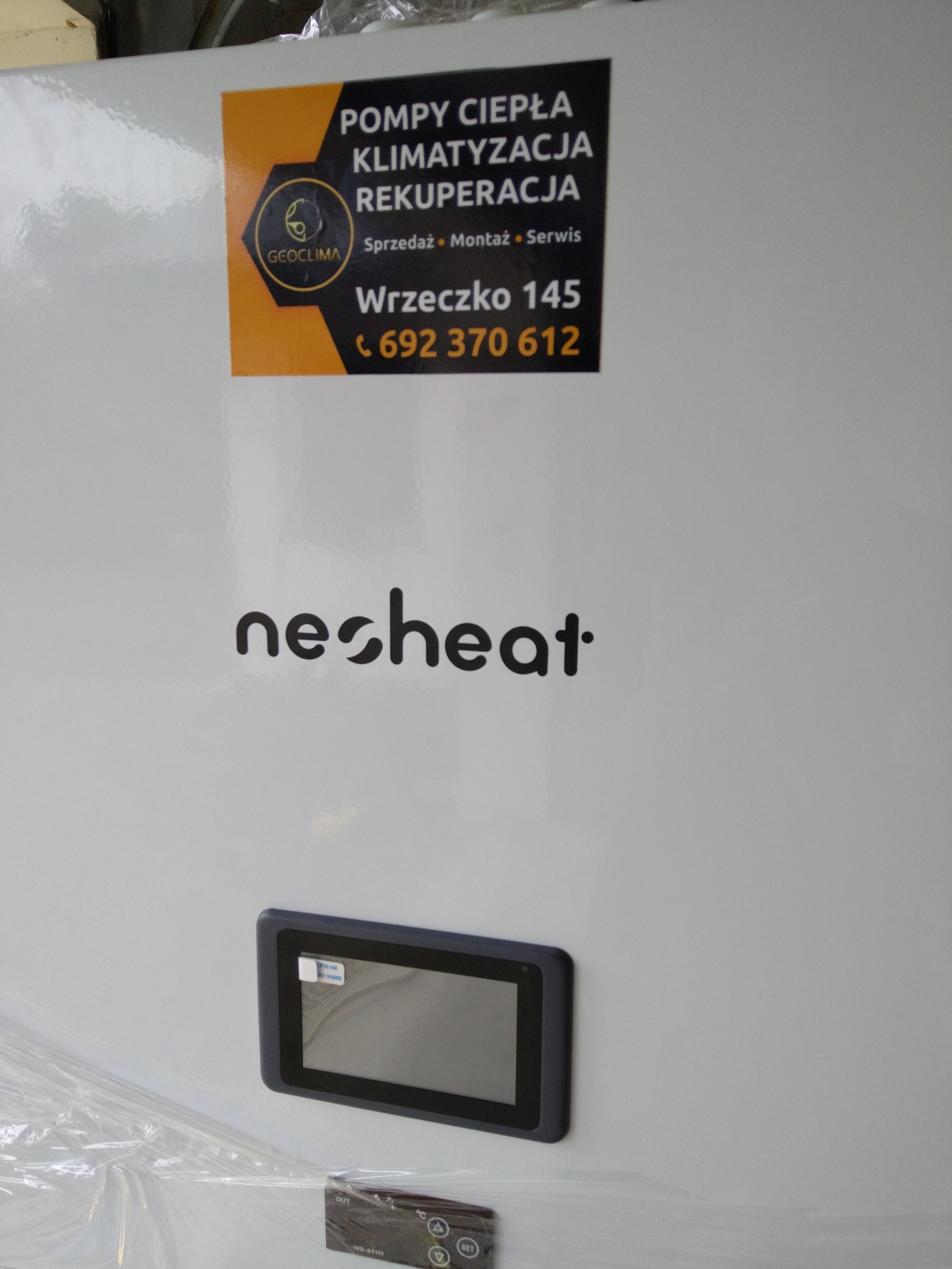 Biała obudowa urządzenia grzewczego z panelem dotykowym i naklejoną wizytówką firmy oferującej sprzedaż, montaż i serwis urządzeń klimatyzacyjnych, rekuperacyjnych i grzewczych, częściowo owinięta...