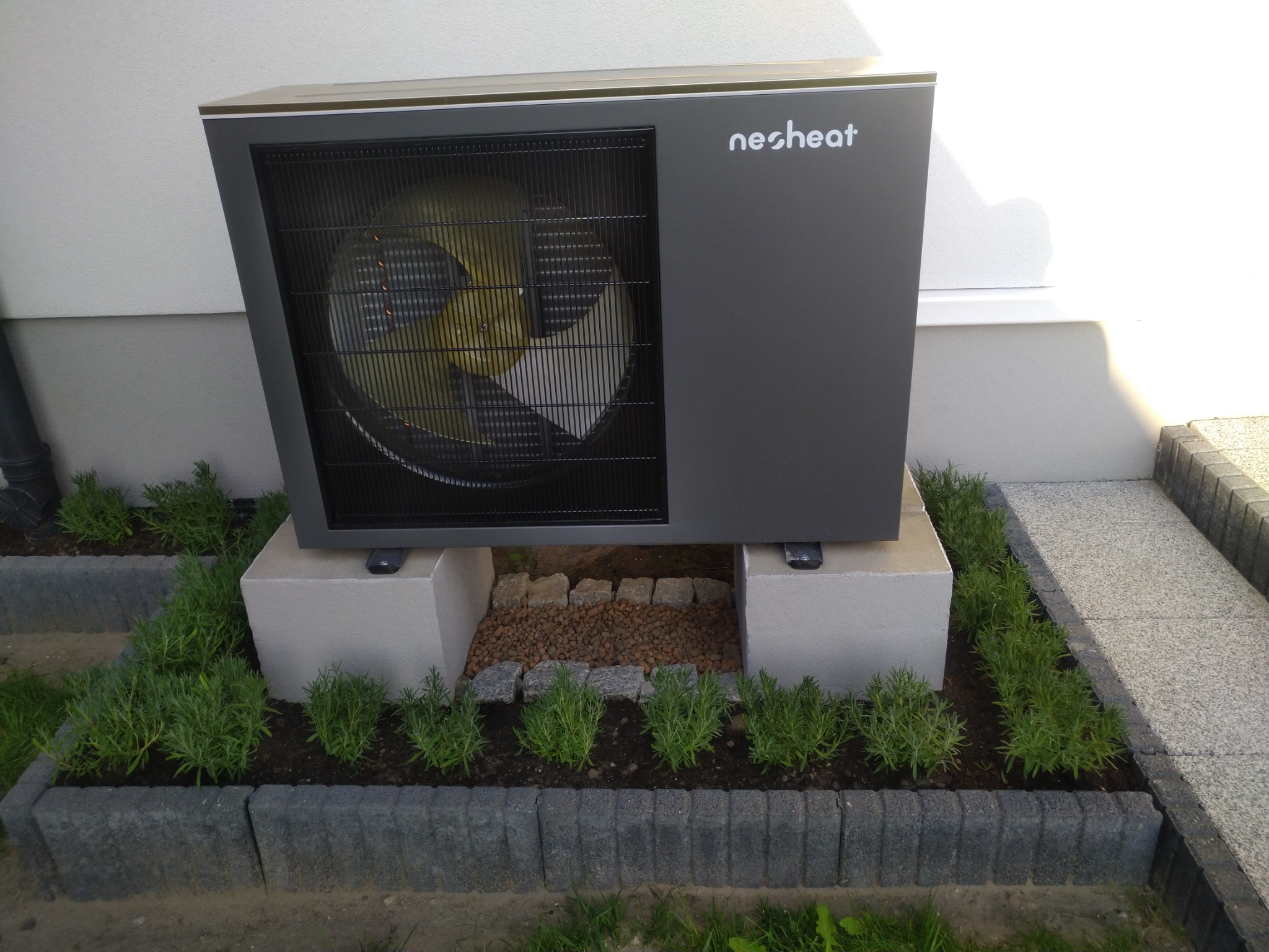 Szara jednostka zewnętrzna z wentylatorem marki nesheat, umieszczona na dwóch betonowych blokach, otoczona rabatą z zielonymi roślinami i kamieniami.
