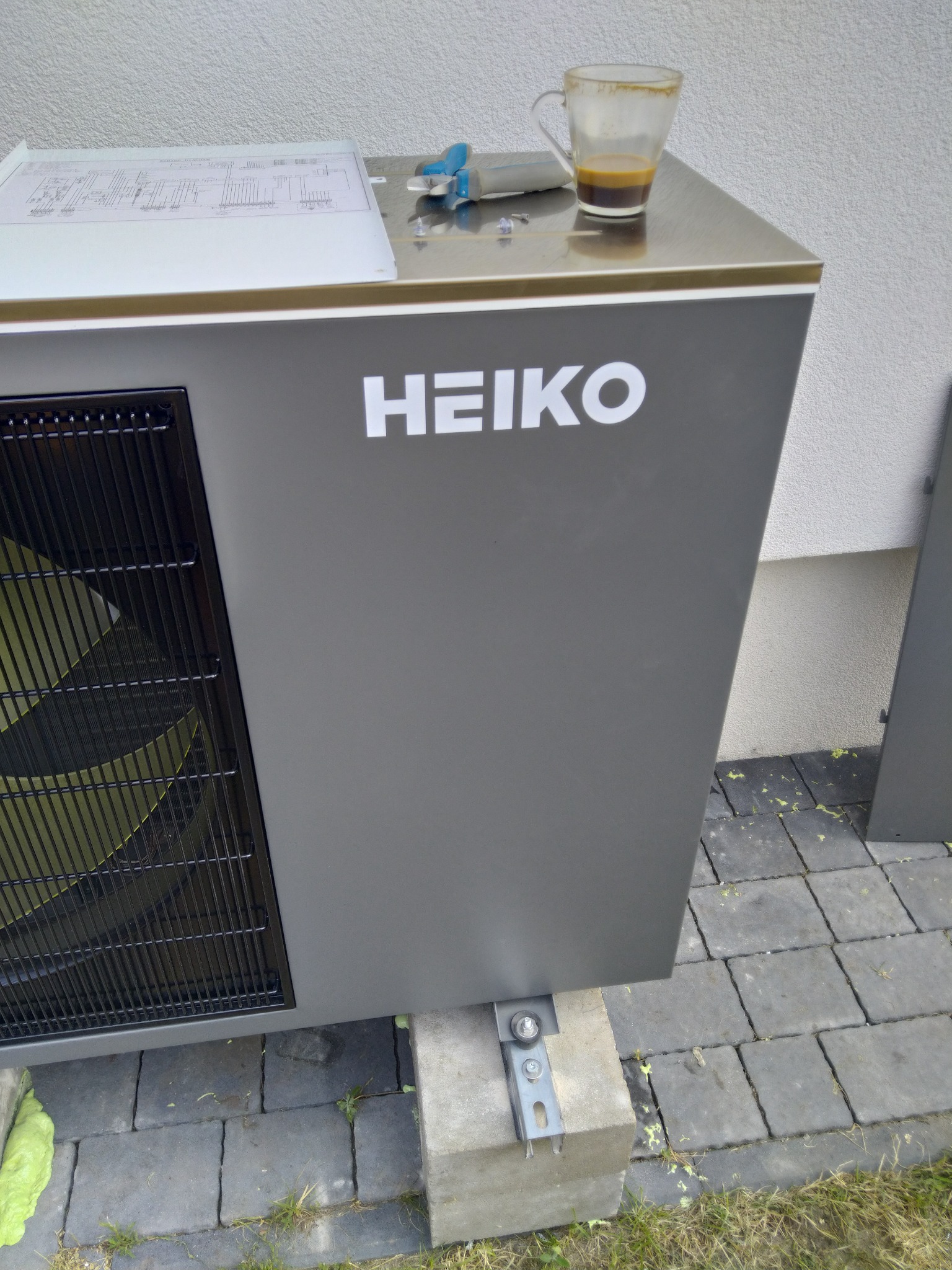 Szara jednostka zewnętrzna z logiem HEIKO, umieszczona na betonowych bloczkach, na górze leży schemat instalacji, obok stoi niedopita kawa w szklance.