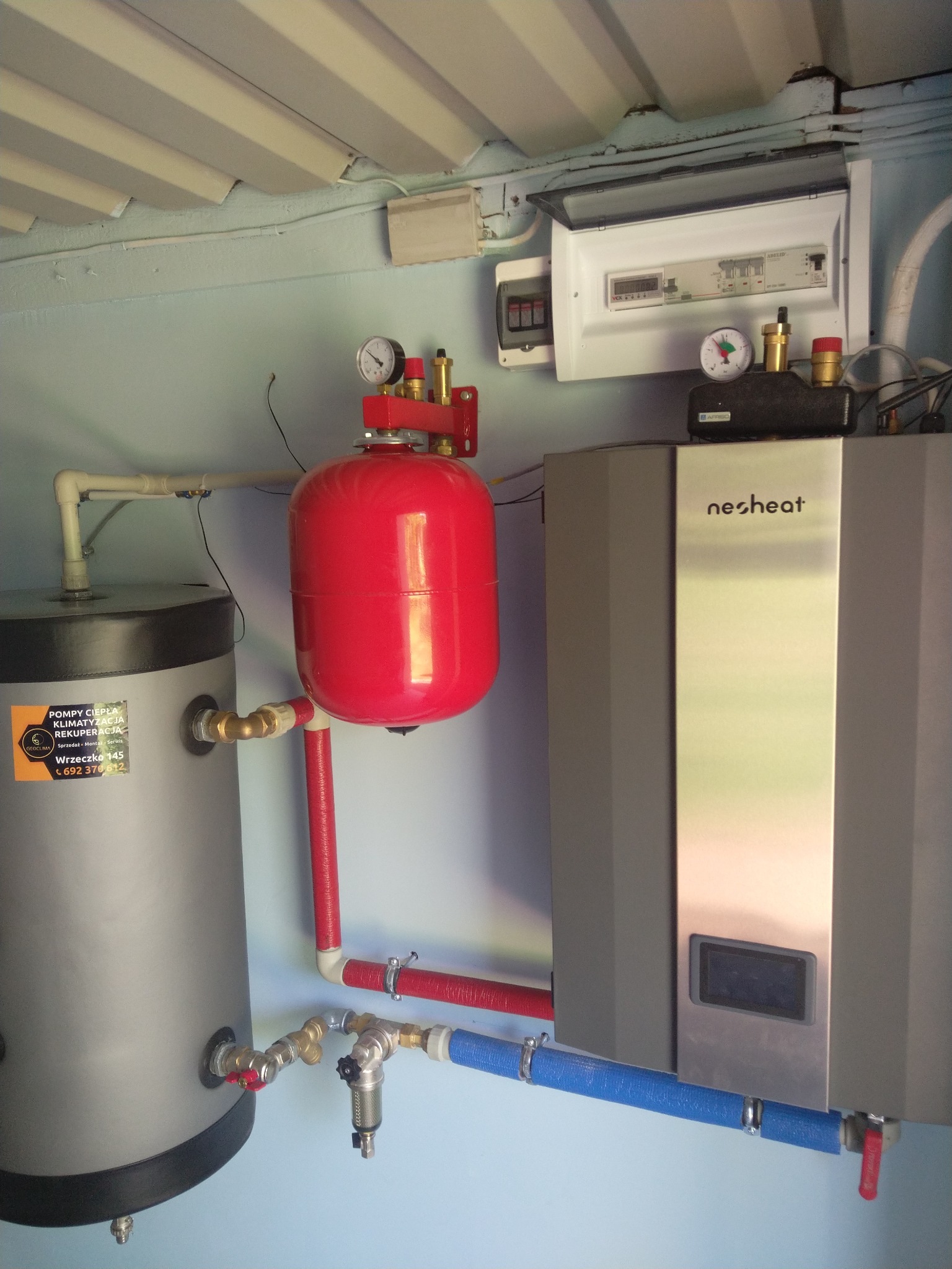 Instalacja grzewcza z szarym zbiornikiem, czerwonym zbiornikiem wyrównawczym i srebrnym urządzeniem grzewczym marki Nesheat, połączone rurami w kolorach czerwonym i niebieskim, zamontowane...