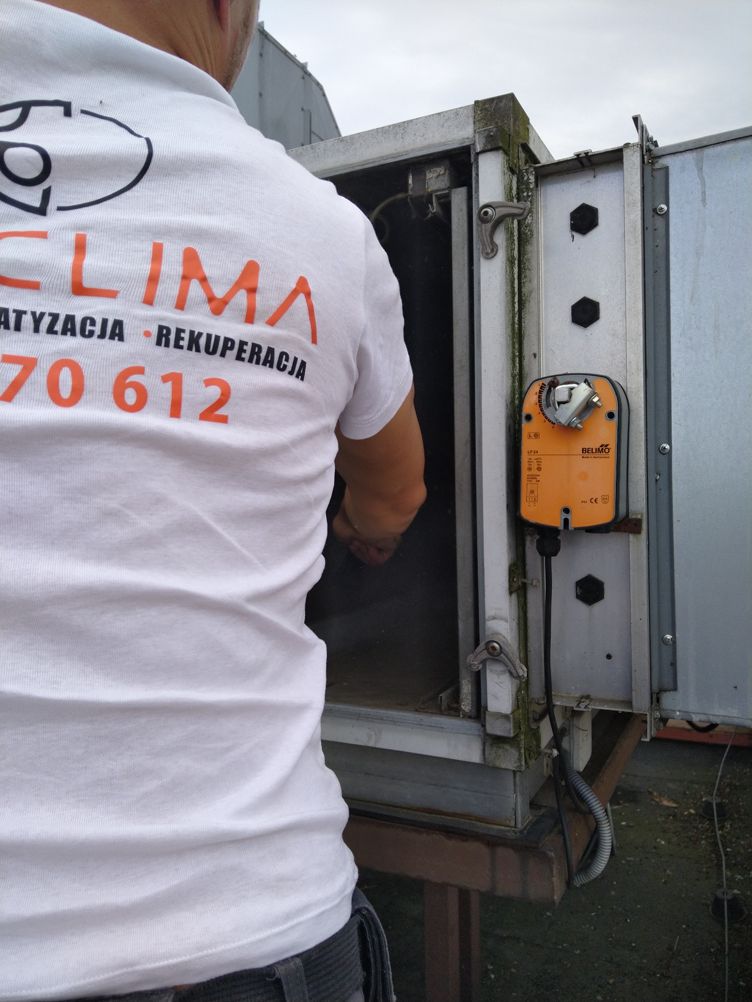 Serwisant w firmowej koszulce z napisem CLIMA, ATYZACJA, REKUPERACJA, 70 612, pracuje przy otwartej, metalowej obudowie urządzenia z pomarańczowym modułem Belimo LF24.