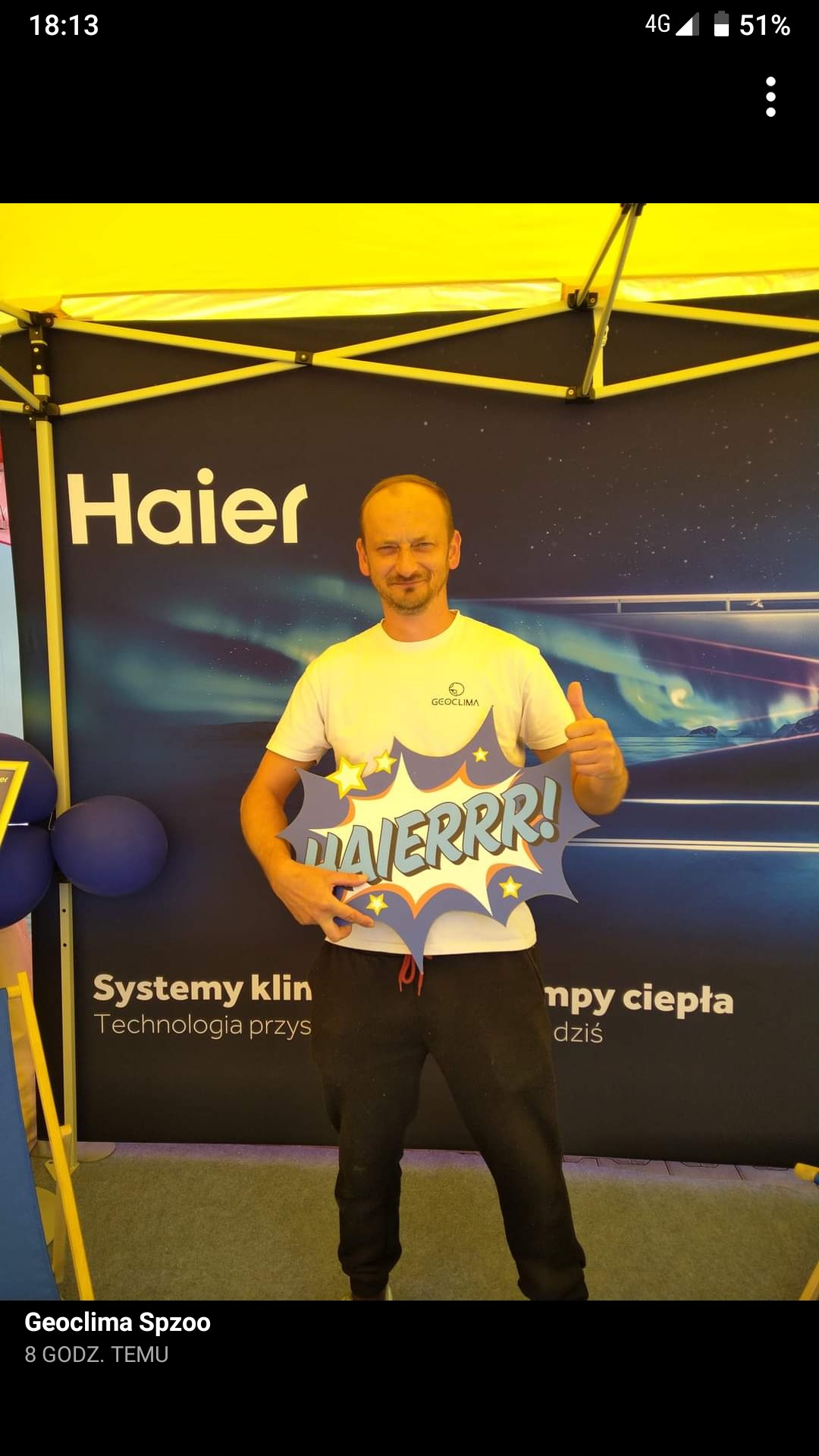 Mężczyzna w białej koszulce z logo Geoclima pozuje z kartonową chmurką z napisem 'HAIERRR!' na tle stoiska z logo Haier i napisem 'Systemy klin... npy ciepła dziś'.