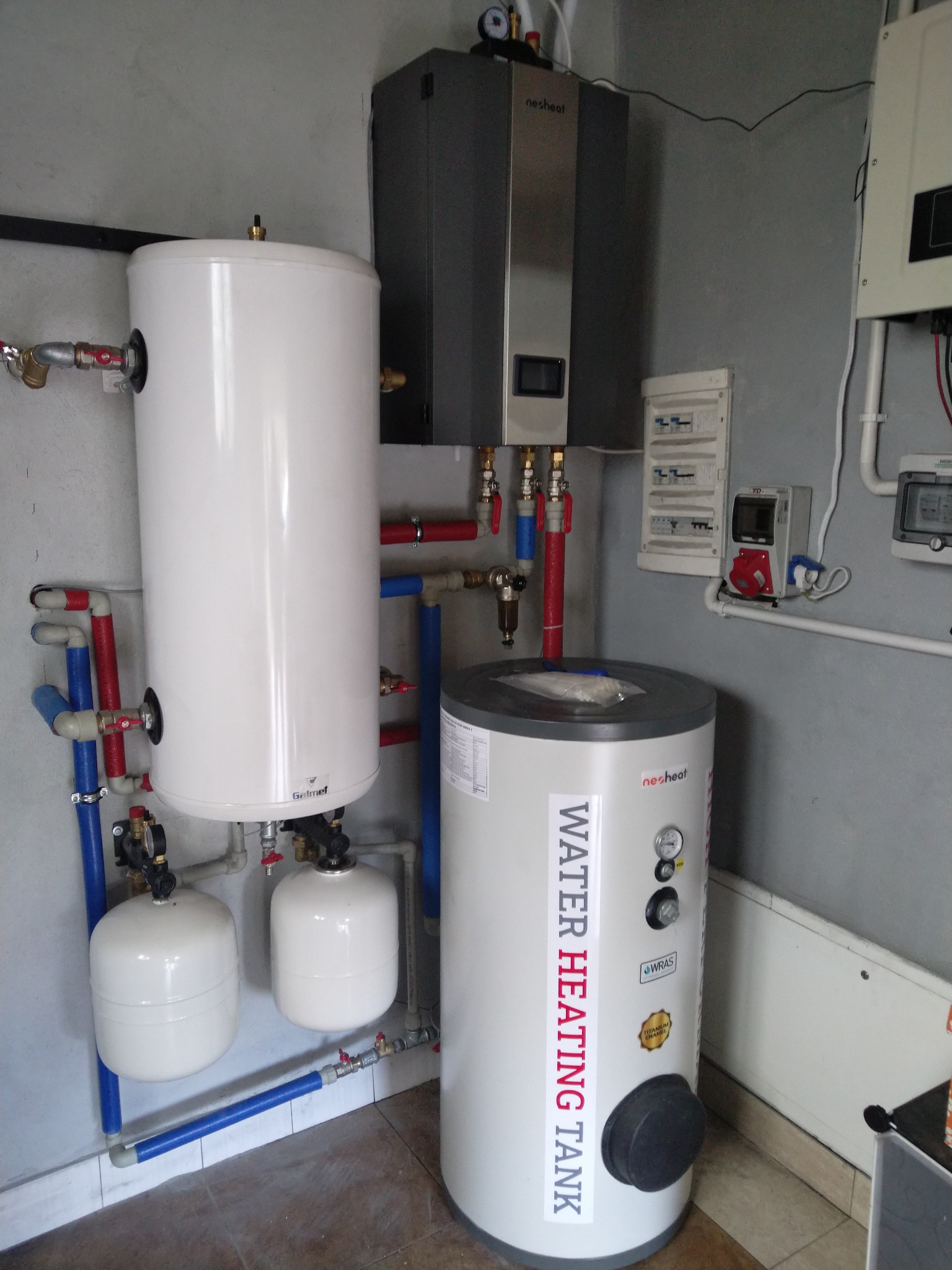 Instalacja pompy ciepła z dwoma zbiornikami: pionowy biały Galmet i szary Nesheat, połączone rurami z elementami hydraulicznymi, na tle szarej ściany z osprzętem elektrycznym.