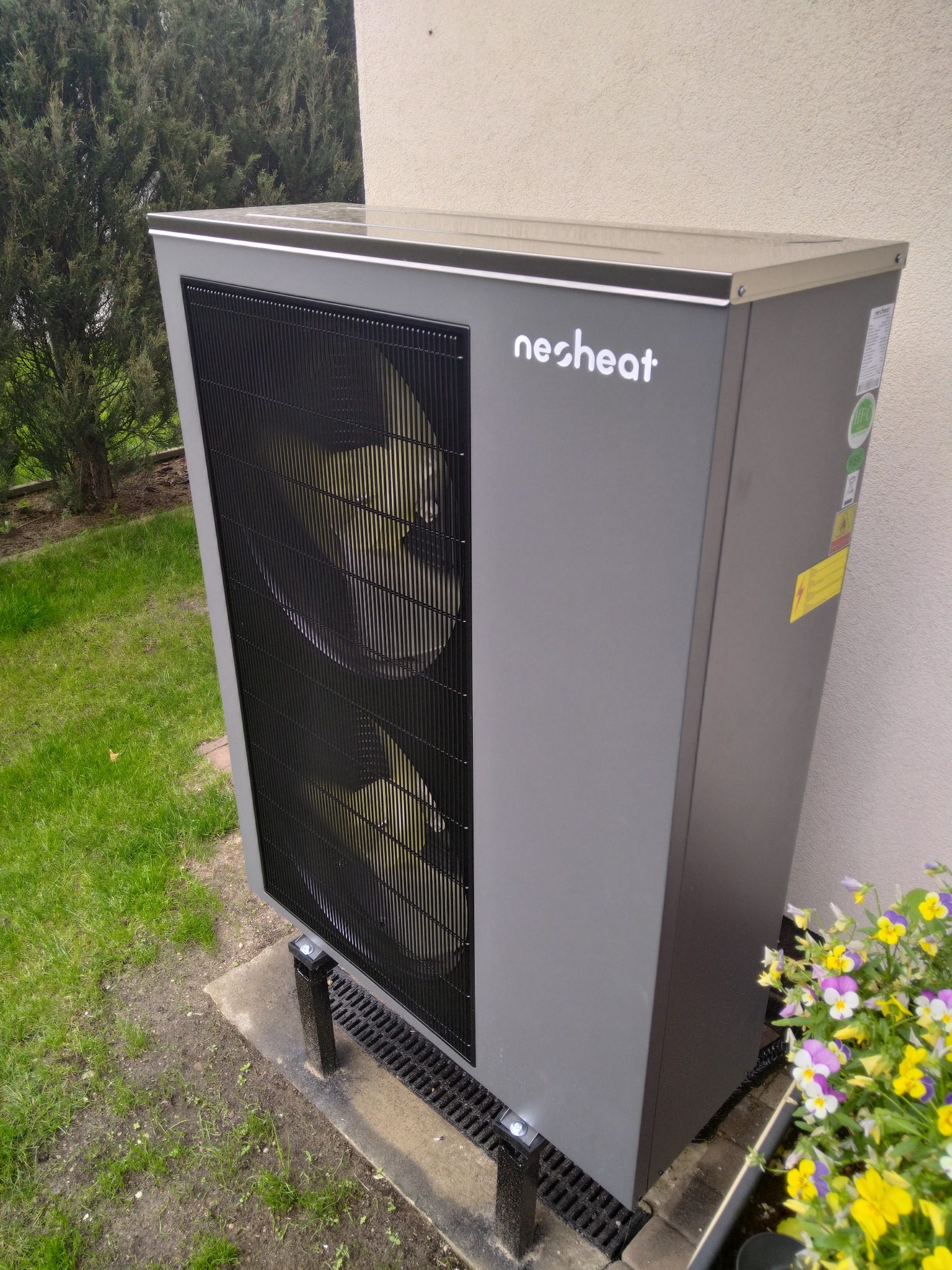Szara pompa ciepła marki necheat zainstalowana na zewnątrz budynku, widoczna na tle zieleni i kwiatów.