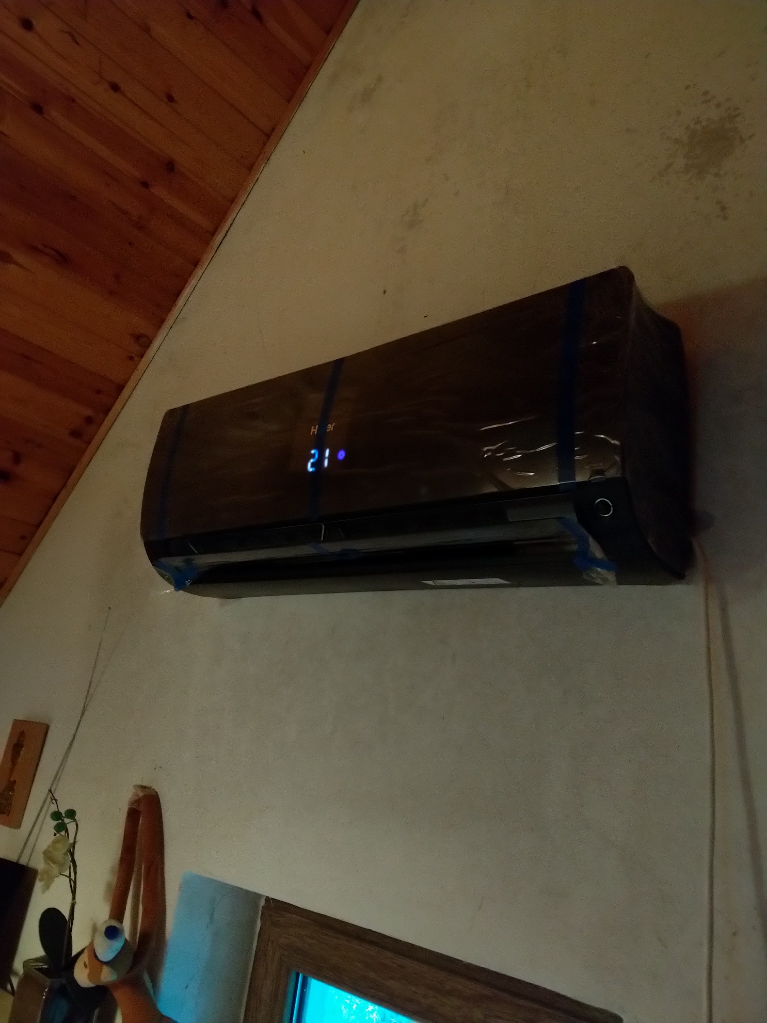 Czarna jednostka klimatyzacji marki Haier, jeszcze w folii ochronnej, zamontowana na ścianie w pomieszczeniu z drewnianym sufitem. Na wyświetlaczu widoczna temperatura 21 stopni.