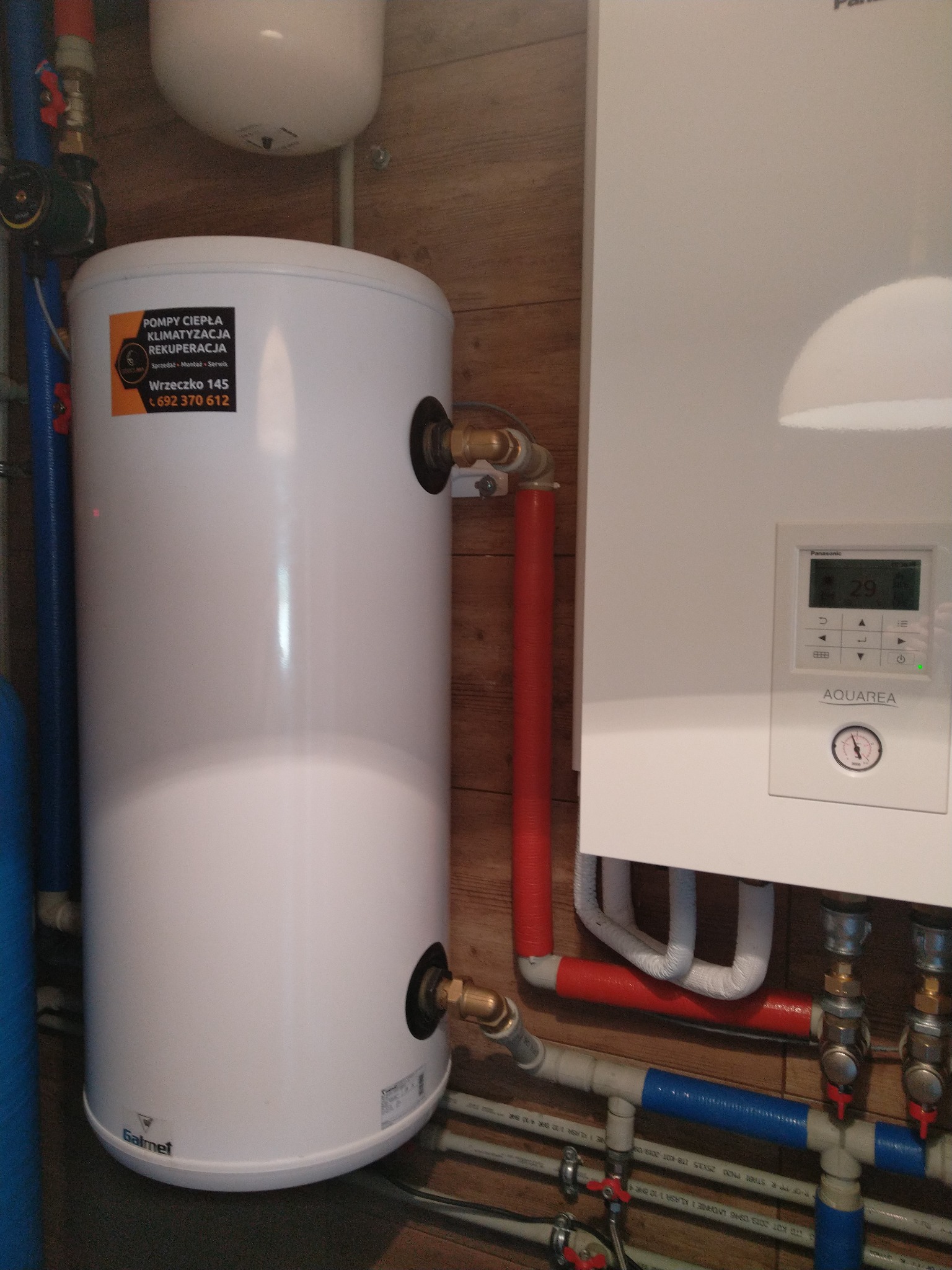 Biały zasobnik ciepłej wody użytkowej Galmet z naklejką informacyjną firmy instalacyjnej, zestawiony z panelem sterującym Panasonic Aquarea i fragmentem instalacji hydraulicznej z rurami w izolacji.