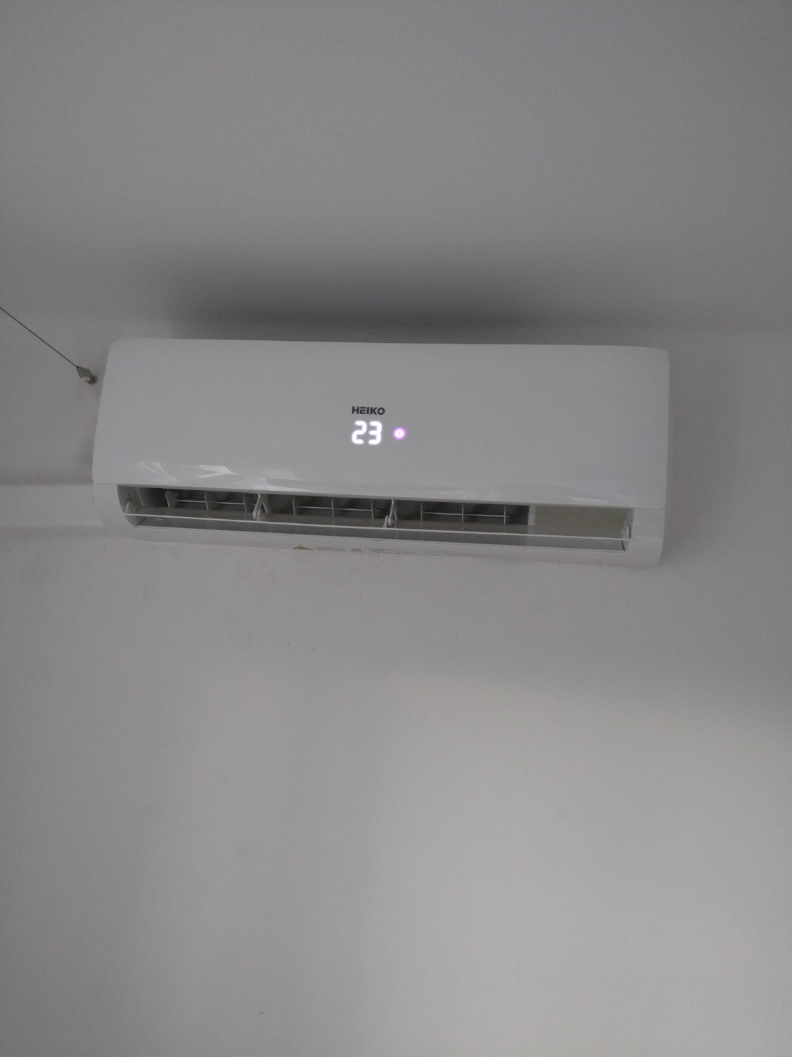 Biały klimatyzator marki HEIKO zamontowany na białej ścianie, wyświetlający temperaturę 23 stopnie Celsjusza.