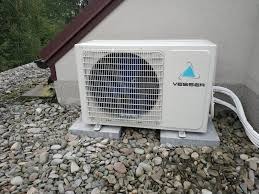 Zewnętrzna jednostka klimatyzacji marki Vesser, zamontowana na betonowych podstawach na żwirowym podłożu, z widocznymi przewodami w białej osłonie.