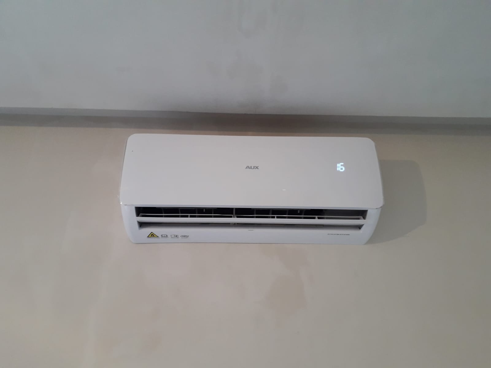 Biała jednostka wewnętrzna klimatyzatora marki AUX zamontowana na jasnej ścianie, wyświetlająca temperaturę 16 stopni Celsjusza.