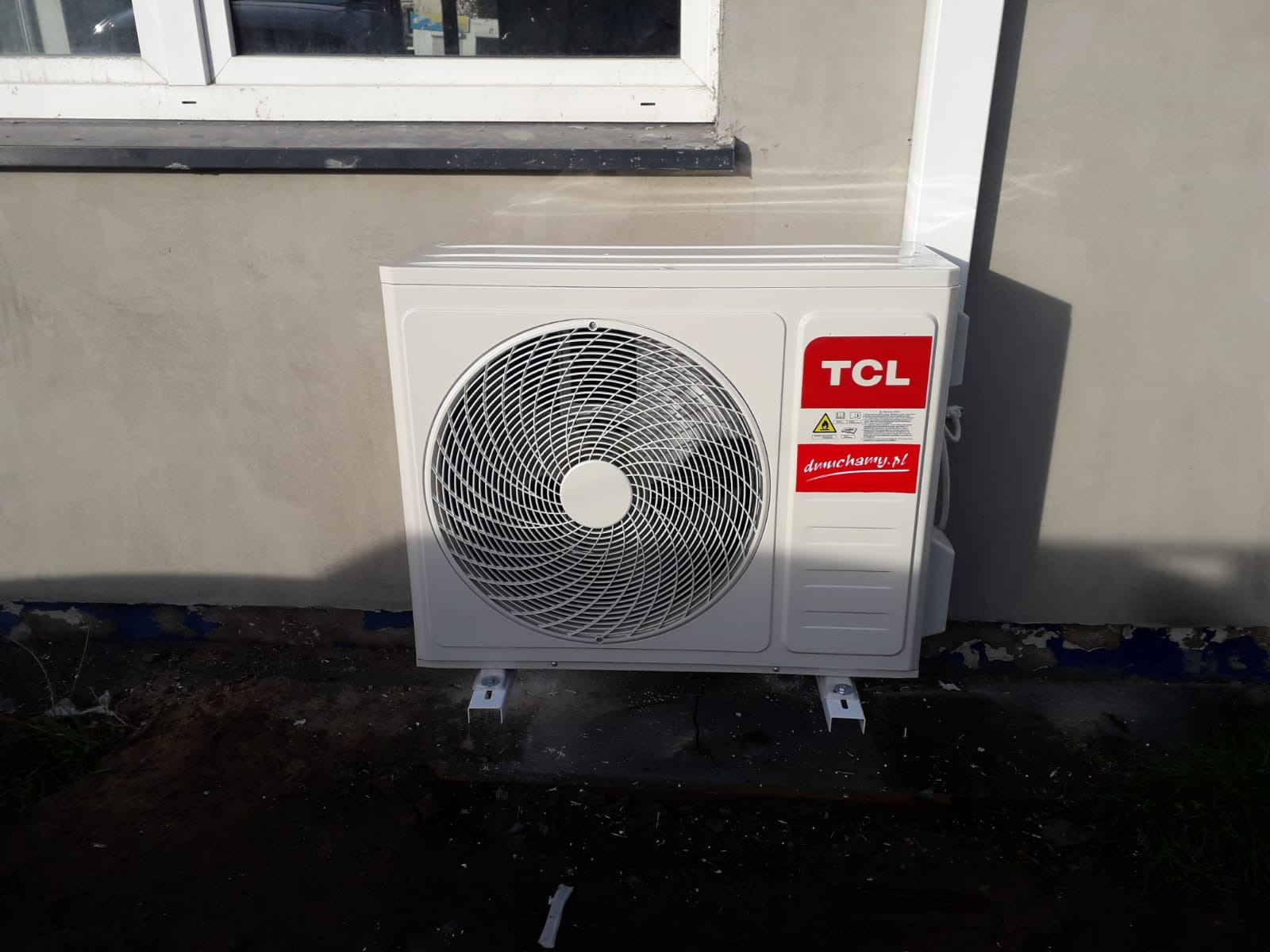 Zewnętrzna jednostka klimatyzacji TCL zamontowana na ścianie budynku, widoczne białe wsporniki montażowe i fragment okna powyżej.