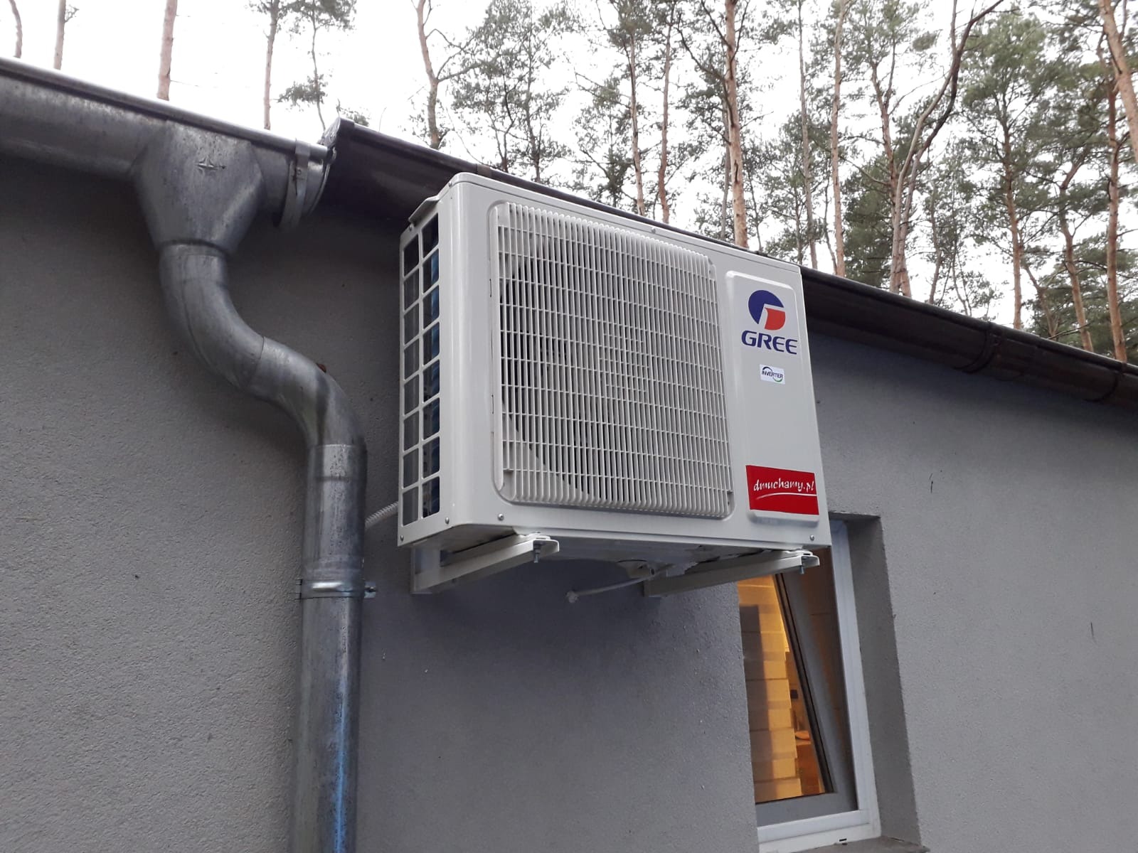 Zewnętrzna jednostka klimatyzacji Gree zamontowana na szarej ścianie budynku z widoczną rynną i oknem, na tle drzew.