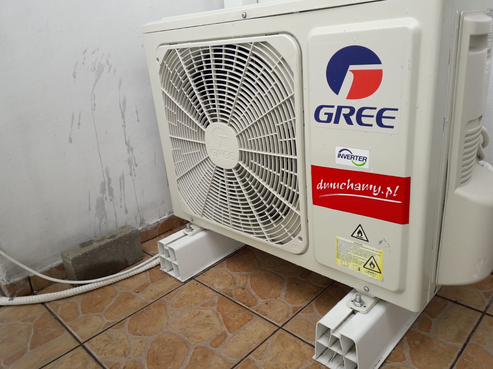 Zewnętrzna jednostka klimatyzacji marki Gree zamontowana na białych, plastikowych wspornikach na brązowej, wzorzystej podłodze, z widocznym przewodem i śladami wilgoci na ścianie.