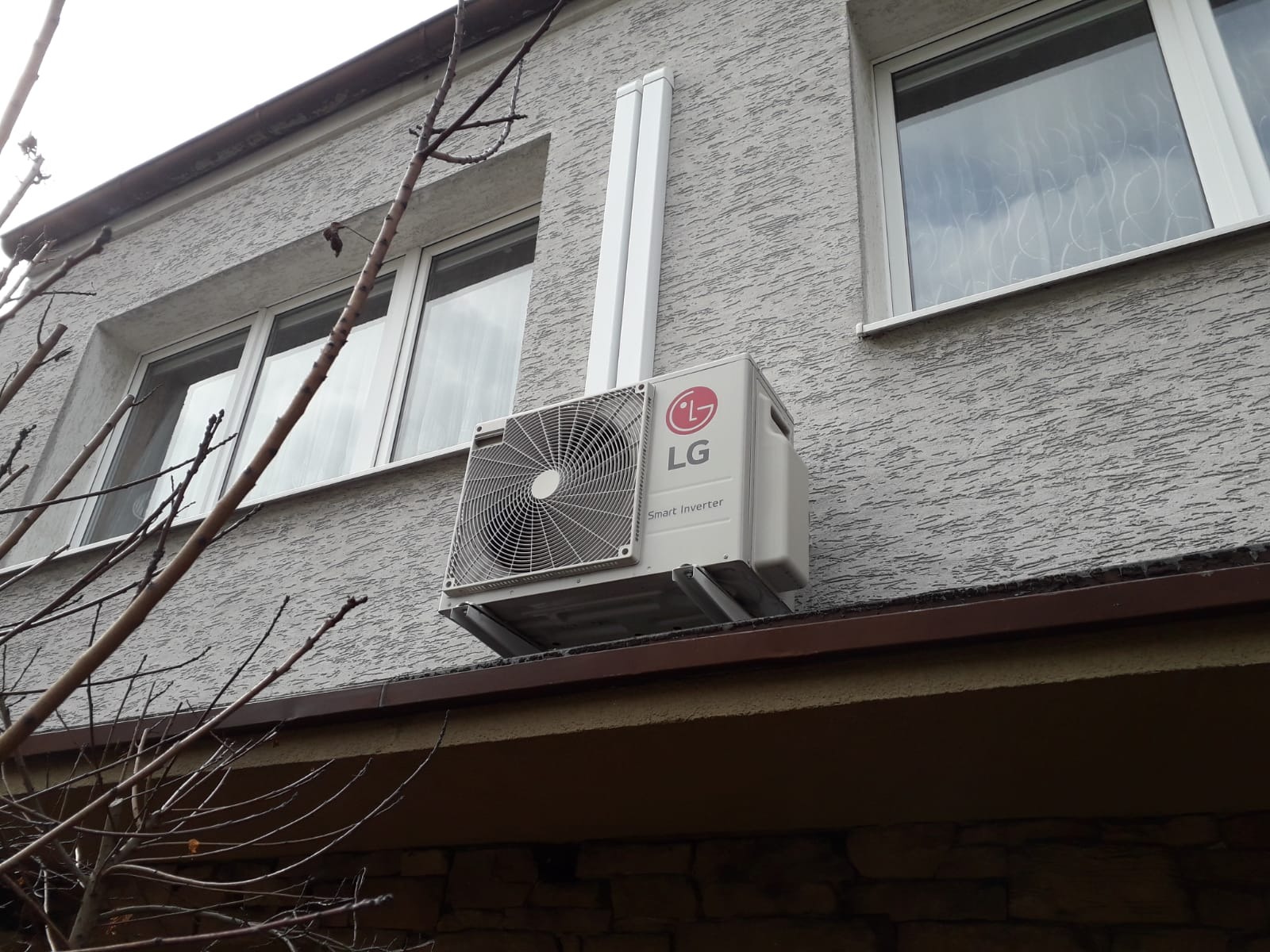 Zewnętrzna jednostka klimatyzatora LG Smart Inverter zamontowana na elewacji domu o szarej strukturze, widoczne orynnowanie i gałęzie drzewa na pierwszym planie.